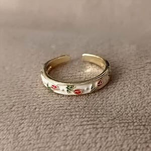 Pode incluir: Um anel aberto de tom dourado com uma incrustação de esmalte branco decorada com um padrão repetitivo de pequenas tulipas vermelhas e folhas verdes. O anel tem um acabamento metálico polido e uma abertura no topo para ajuste de tamanho.