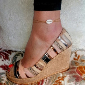 May include: A gold chain anklet with a white seashell charm. The anklet is worn on a person's ankle, and the seashell is centered on the ankle.