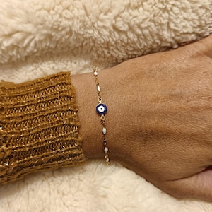 Peut inclure: Bracelet doré orné d'un charm en forme d'œil bleu et blanc. La chaîne délicate est ornée d'accents blancs ovales. Le bracelet est porté sur un poignet avec une manche de pull dorée.