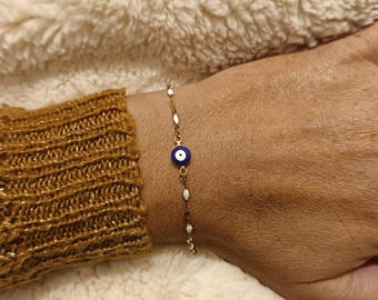 Può includere: Bracciale dorato con un ciondolo a forma di occhio blu e bianco. Il bracciale ha una catena delicata con accenti ovali bianchi. Il bracciale è indossato su un polso con una manica di maglione dorata.