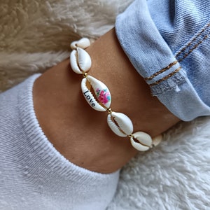 Könnte beinhalten: Ein Fußkettchen mit weißen Kaurimuscheln, die mit kleinen goldfarbenen Perlen aufgereiht sind. Eine Muschel ist mit einem rosa Blumenmuster und dem Wort Love in schwarzen Buchstaben verziert. Die Muscheln bilden eine durchgehende kreisförmige Kette.