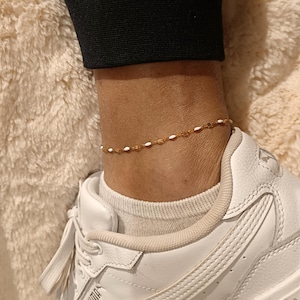 Peut inclure: Un bracelet de cheville doré délicat orné de petites perles blanches et ambrées. Le bracelet est porté sur une cheville, avec une basket blanche visible en dessous. Le fond est une surface douce et texturée.