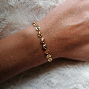 Könnte beinhalten: Ein goldenes Armband mit kleinen runden Anhängern, die jeweils einen bunten Mittelpunkt haben. Das Armband wird am Handgelenk getragen.