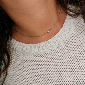 Peut inclure: Un collier fin en chaîne dorée avec un petit pendentif bleu en forme d'œil maléfique. Le collier est délicat et fin.