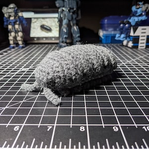 May include: A gray crocheted plush toy shaped like a pill bug. The toy is sitting on a black gridded surface.