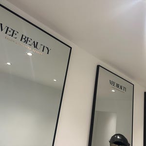 May include: Three large mirrors with black frames are mounted on a white wall. The mirrors display the text "VEE BEAUTY SALON & TRAINING ACADEMY" in a black, elegant font. The mirrors are evenly spaced and lit by recessed ceiling lights.