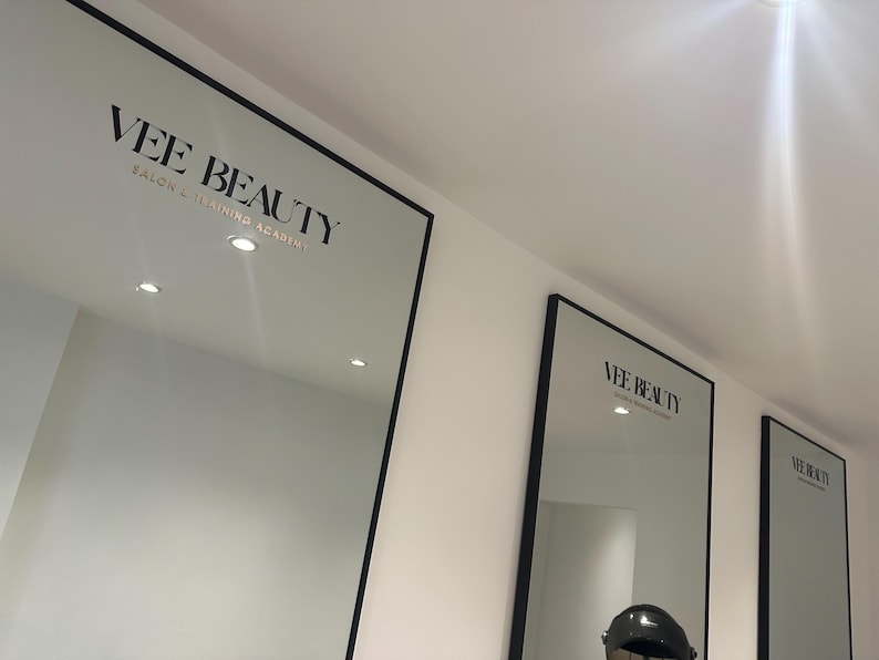 May include: Three large mirrors with black frames are mounted on a white wall. The mirrors display the text "VEE BEAUTY SALON & TRAINING ACADEMY" in a black, elegant font. The mirrors are evenly spaced and lit by recessed ceiling lights.