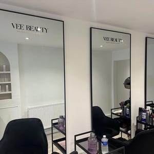 May include: Three large, rectangular mirrors with black frames are mounted on a white wall. The mirrors reflect a salon interior with black chairs, shelves, and beauty products. The text "VEE BEAUTY" is displayed at the top of each mirror.