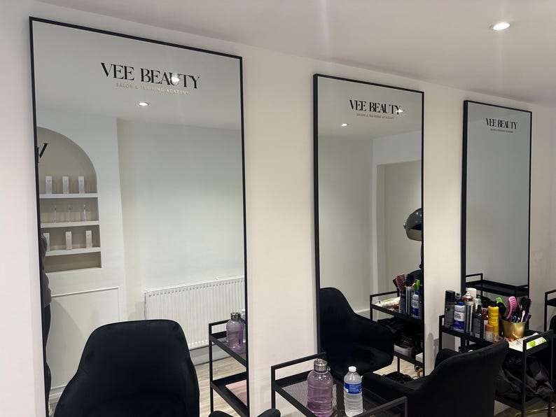 May include: Three large, rectangular mirrors with black frames are mounted on a white wall. The mirrors reflect a salon interior with black chairs, shelves, and beauty products. The text "VEE BEAUTY" is displayed at the top of each mirror.