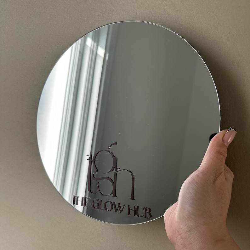 May include: A round, silver mirror with the text "tgh THE GLOW HUB" in a dark brown font. The mirror is being held by a hand with a manicure. The mirror reflects the surrounding environment.