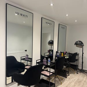 May include: Interior shot of a beauty salon with three large mirrors with black frames. Each mirror has the text "Vee Beauty" at the top. Black chairs with gold bases are in front of the mirrors, along with black rolling carts.