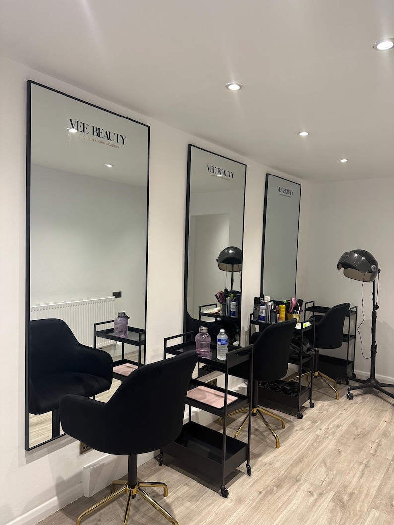 May include: Interior shot of a beauty salon with three large mirrors with black frames. Each mirror has the text "Vee Beauty" at the top. Black chairs with gold bases are in front of the mirrors, along with black rolling carts.