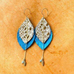 May include: A pair of dangle earrings with a layered design. The top layer is a silver and gold faux leather feather, the middle layer is a blue faux leather feather, and the bottom layer is a gold chain with a small gold feather charm.