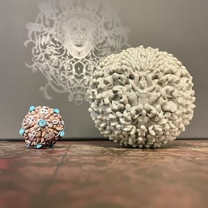 Może przedstawiać: Dwie dekoracyjne kule, jedna duża i jedna mała, wykonane z białej ceramiki. Większa kula jest pokryta małymi figurkami, podczas gdy mniejsza kula jest pokryta mniejszymi, bardziej szczegółowymi figurkami. Kule znajdują się na drewnianej powierzchni.