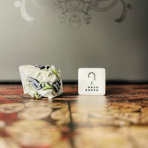 Puede incluir: Dos dados blancos, uno con un diseño detallado y el otro con un signo de interrogación y puntos. Los dados están sobre una superficie de madera con un fondo borroso.