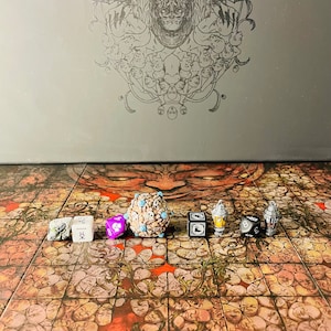 Op de afbeelding: Een verzameling dobbelstenen en miniatuurspellen gerangschikt op een spelbord. De dobbelstenen zijn in verschillende kleuren en stijlen, waaronder een paarse D8, een zwarte D6 en een witte D6. Het spelbord heeft een herhalend gezichtspatroon.