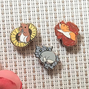 Pode incluir: Três pinos de esmalte com desenhos de animais fofinhos. O primeiro pino mostra um hamster marrom em um girassol amarelo. O segundo pino mostra um hamster branco e laranja em uma rosa vermelha. O terceiro pino mostra um hamster cinza em uma flor roxa.