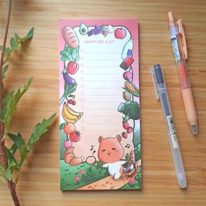 May include: A rectangular shopping list notepad with a pink gradient background. The notepad features a cartoon illustration of a hamster and various fruits and vegetables. The words "SHOPPING LIST" are printed at the top. Two pens are next to the notepad.