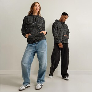 May include: Two people wearing matching black camouflage hoodies with a light brown outline design. One person is wearing light blue jeans, and the other is wearing black cargo trousers. Both are wearing white trainers with black stripes.