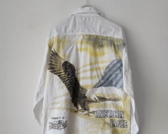 May include: White long-sleeve button-up shirt with a faded American Eagle graphic on the back. The graphic features a bald eagle flying over a mountain range with a blue and white striped flag in the background. The shirt also has the text "PRESENT BY" and "STOCKERPOLE" on the left sleeve.