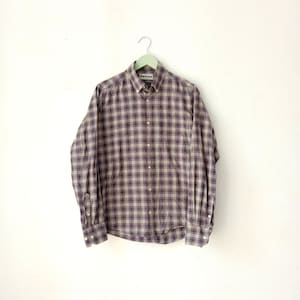 Puede incluir: Una camisa de manga larga a cuadros con un patrón de cuadros marrones y morados. La camisa tiene un cuello abotonado y una etiqueta de Barbour en el pecho izquierdo.