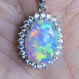 May include: A silver necklace featuring an oval opal pendant surrounded by a halo of small, clear gemstones. The opal displays a play of color with hues of blue, green, and yellow. The chain is a delicate silver link.