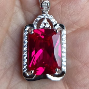 May include: A silver pendant necklace featuring a large, rectangular, vibrant red gemstone. The gemstone is set in a silver frame with a halo of small, clear stones. The necklace has a delicate silver chain.