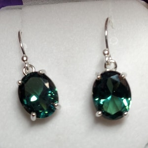 May include: A pair of silver dangle earrings with oval-shaped green gemstones. The earrings are displayed in a purple seashell-shaped box.