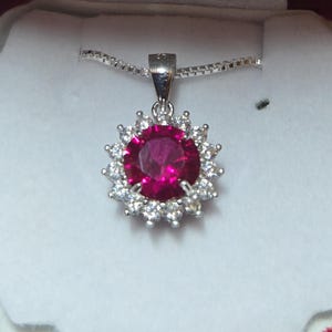 May include: A silver pendant necklace featuring a large, round, ruby-red gemstone surrounded by smaller, clear gemstones. The pendant is suspended from a delicate silver chain, presented in a white box with a red trim.