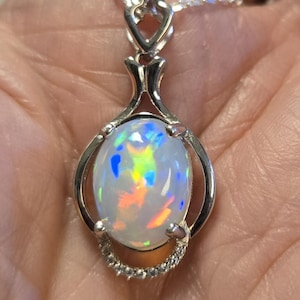 May include: A silver pendant necklace featuring a central oval-shaped opal gemstone with iridescent flashes of blue, green, yellow, and orange. The stone is set in a polished silver frame with a small decorative arch at the top and a chain.