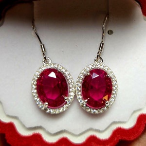 May include: A pair of silver dangle earrings with a large oval-shaped red gemstone surrounded by a halo of smaller white gemstones.