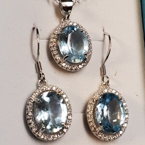 May include: A silver-toned jewelry set featuring a pendant necklace and matching earrings. Each piece showcases a large, oval, light blue gemstone surrounded by a halo of small, clear stones. The set is displayed on a light gray surface.