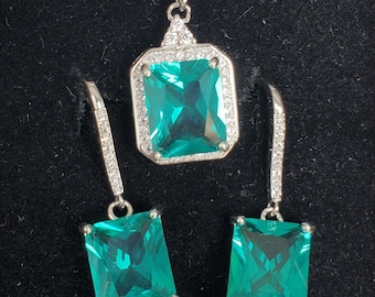 May include: A silver necklace and earrings set with a large, square, emerald green gemstone surrounded by smaller clear gemstones.