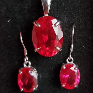 May include: A silver necklace and earrings set with red oval-shaped gemstones. The necklace has a delicate chain and a pendant with a large red gemstone. The earrings are simple studs with smaller red gemstones.