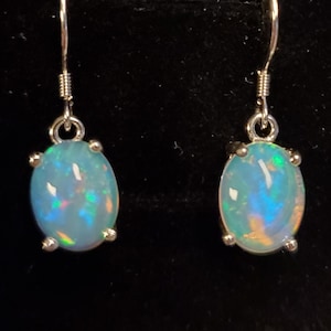 Pu&ograve; includere: Un paio di orecchini pendenti con opali ovali e iridescenti incastonati in griffe di metallo color argento. Ogni orecchino pende da un semplice gancio color argento, mostrando una base bianco latte con bagliori di luce blu, verde e arancione.