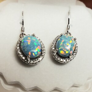 May include: A pair of silver dangle earrings with oval-shaped opal gemstones surrounded by a halo of smaller clear stones.