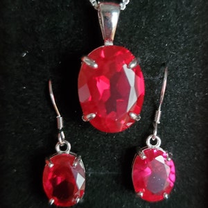 Puede incluir: Un collar de plata con un colgante ovalado rojo y pendientes a juego. El colgante y los pendientes presentan una gran piedra roja facetada.
