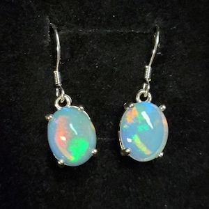 May include: Pair of elegant sterling silver opal earrings. Each earring features a light blue oval-shaped opal gemstone with vibrant internal flashes of orange, green, and blue. The opals are securely set in silver claw settings, adding to their refined look. These dangle earrings are perfect for adding a touch of sophistication to any outfit.