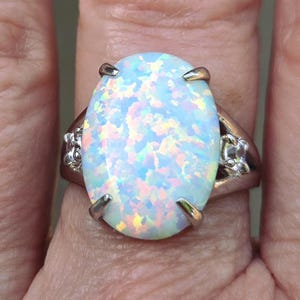May include: A silver ring featuring a large, oval opal gemstone. The opal displays a vibrant play-of-color with flashes of blue, green, and yellow. The ring's setting has decorative details on the sides, and the opal is held in place by four prongs.