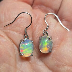 Pu&ograve; includere: Un paio di orecchini pendenti color argento con opali di forma ovale. Ogni opale mostra un gioco di colori con riflessi verdi, gialli e arancioni su una base bianca traslucida. Le pietre sono fissate in montature a quattro griffe con ganci.