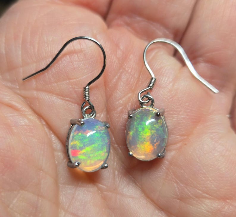 Pu&ograve; includere: Un paio di orecchini pendenti color argento con opali di forma ovale. Ogni opale mostra un gioco di colori con riflessi verdi, gialli e arancioni su una base bianca traslucida. Le pietre sono fissate in montature a quattro griffe con ganci.