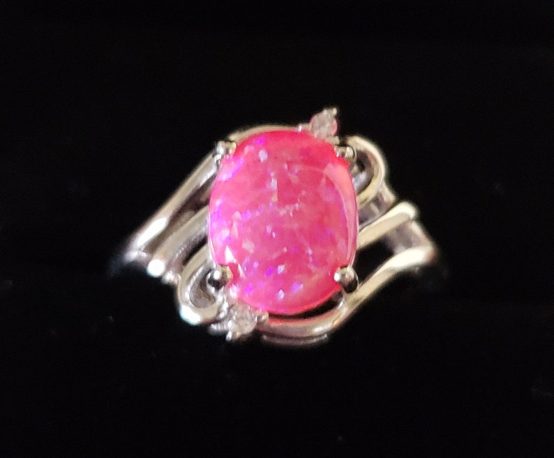 May include: A silver ring with a pink opal gemstone set in a swirling design. The ring has small clear accent stones.