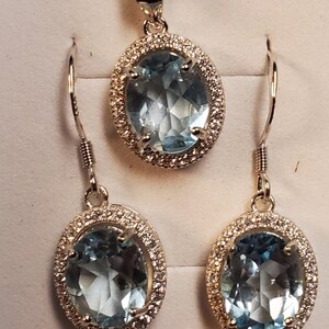 May include: A jewelry set featuring a pendant necklace and matching earrings. The set includes an oval-shaped, light blue gemstone pendant and earrings, each surrounded by a halo of small, clear stones. The jewelry is set in a silver-colored metal.