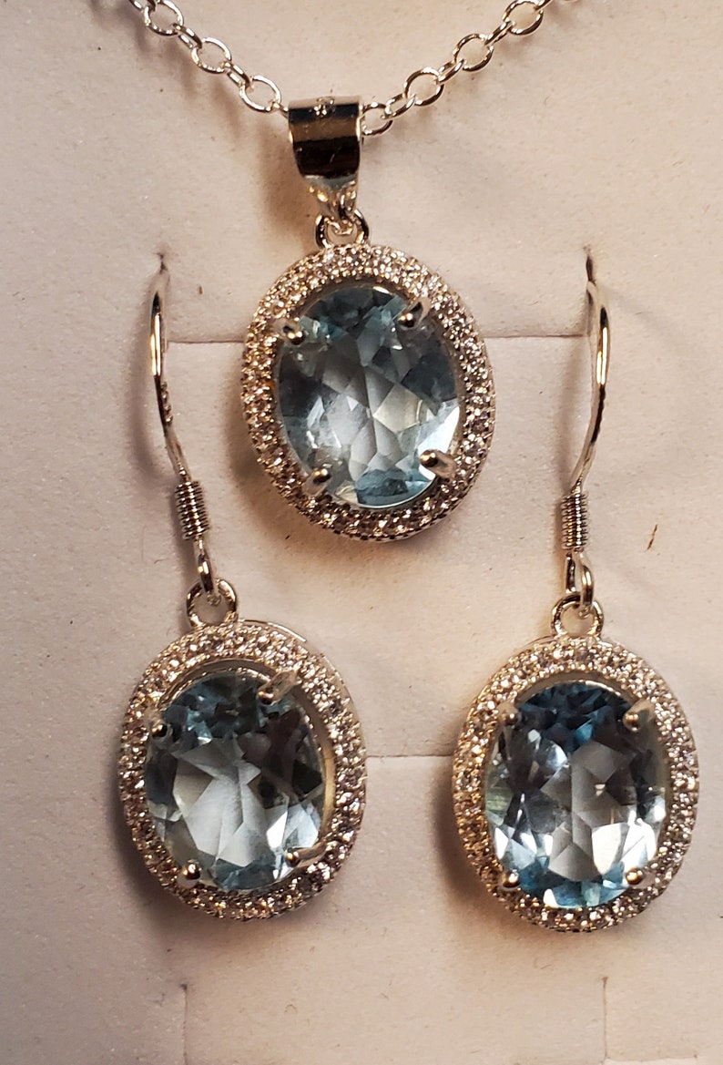 May include: A jewelry set featuring a pendant necklace and matching earrings. The set includes an oval-shaped, light blue gemstone pendant and earrings, each surrounded by a halo of small, clear stones. The jewelry is set in a silver-colored metal.