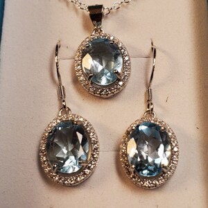 May include: A silver-toned jewelry set featuring a necklace pendant and matching earrings. Each piece has an oval, light blue gemstone surrounded by a halo of small, clear stones. The necklace has a delicate chain.