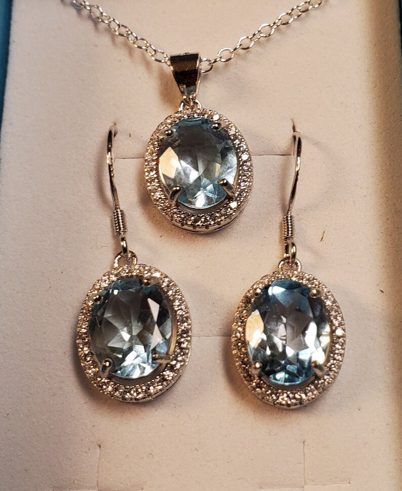 May include: A silver-toned jewelry set featuring a necklace pendant and matching earrings. Each piece has an oval, light blue gemstone surrounded by a halo of small, clear stones. The necklace has a delicate chain.