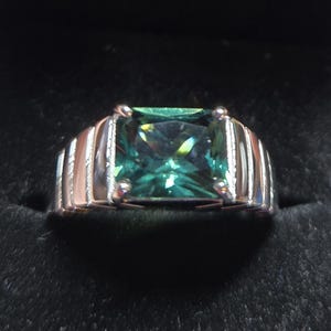 May include: A silver ring with a large, rectangular, teal-colored gemstone. The ring's band has a ribbed design on the sides. The gemstone is set in a four-prong setting, and the ring is displayed against a black background.