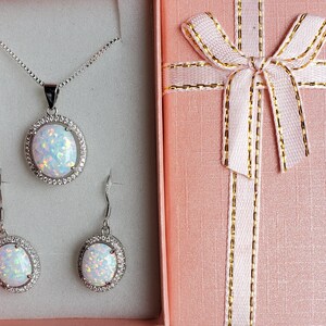 May include: A white opal necklace and earrings set in a silver gift box. The necklace features a large oval opal pendant with a silver halo. The earrings are also oval shaped with silver halos.