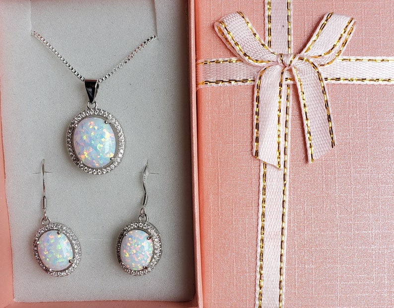 May include: A white opal necklace and earrings set in a silver gift box. The necklace features a large oval opal pendant with a silver halo. The earrings are also oval shaped with silver halos.