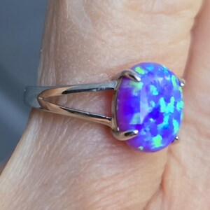 May include: A silver-toned ring featuring an oval-shaped, iridescent purple and blue opal-style stone set in a four-prong mounting. The band has a split-shank design that tapers toward the back, showcasing a polished metallic finish throughout.